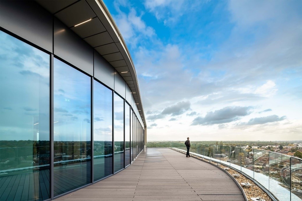 Modern building with curved glass facade and wooden deck overlooking trees and distant buildings.