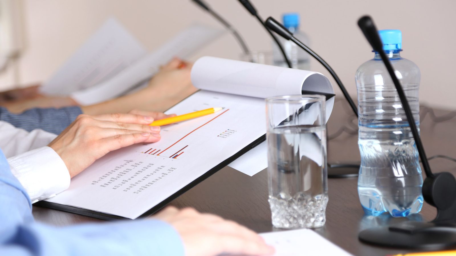 A person writing notes on a piece of paper and seated a table with meeting microphones.