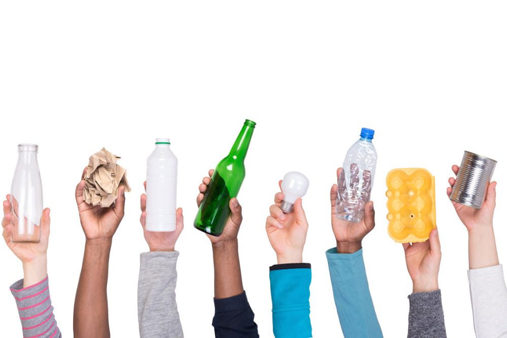 Image of multiple hands holding up different types of recycable waste including glass bottles, plastic bottles and cans