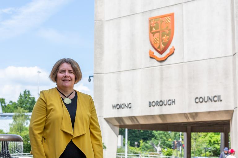 Cllr Ann-Marie Barker wearing a yellow gold jacket standing in front of the Woking Borough Council offices