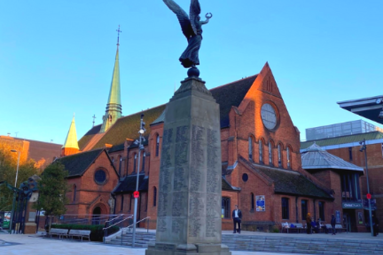 Stone war memorial with engraved names and a bronze winged figure on top, set in a paved square with a grass base and red poppy wreaths. A red-brick church with a tall spire is behind it under a clear blue sky.