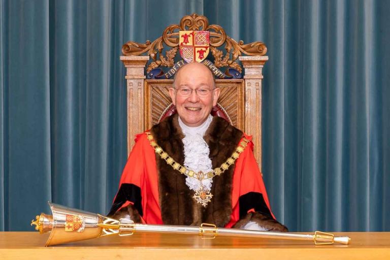 Cllr Rob Leach wearing the Mayor of Woking's ceremonial clothes, including a red cape, large gold chain and ruffled white caveat. He is sitting in front of the Mayor's gold mace which is laid flat on a wooden desk.