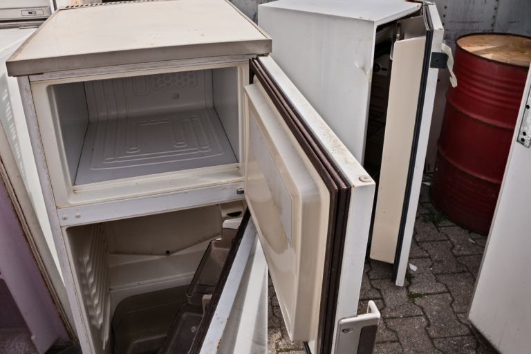 A broken fridge with an open door with other old fridges and freezers in the background.
