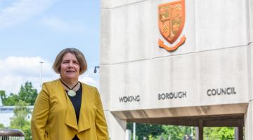 Cllr Ann-Marie Barker wearing a yellow gold jacket standing in front of the Woking Borough Council offices