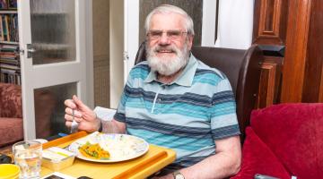 Man with grey hair and beard sitting in a chair eating a meal in his home