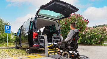 Man in wheelchair behind an accessible taxi with its boot open.
