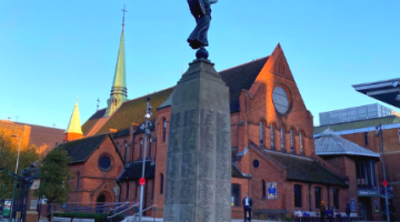 Stone war memorial with engraved names and a bronze winged figure on top, set in a paved square with a grass base and red poppy wreaths. A red-brick church with a tall spire is behind it under a clear blue sky.