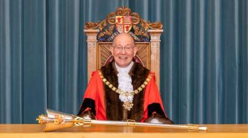 Cllr Rob Leach wearing the Mayor of Woking's ceremonial clothes, including a red cape, large gold chain and ruffled white caveat. He is sitting in front of the Mayor's gold mace which is laid flat on a wooden desk.