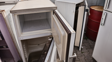 A broken fridge with an open door with other old fridges and freezers in the background.